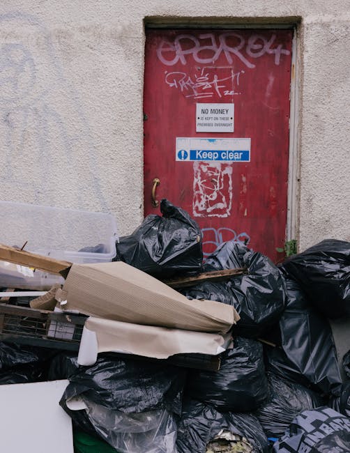 A collection of black rubbish bags and discarded packaging materials piled in front of a red metal door set into a beige, textured concrete wall. The bags appear full and are stacked haphazardly, with some leaning against the wall and others resting on cardboard and plastic packaging. The red door has multiple graffiti tags in white and pink, along with a white sign that reads 'NO MONEY' and 'KEEP CLEAR' in blue, with a small padlock handle visible on the left side. The surrounding environment suggests an outdoor urban location, possibly an alleyway or service area where waste is temporarily stored before collection, highlighting the context of private or alternative waste disposal methods a company like Rubbish Collection Barnet might facilitate.