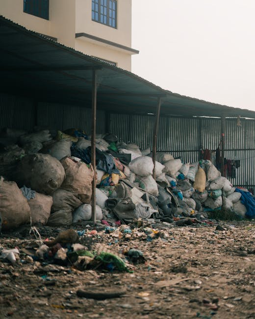 A large accumulation of black and transparent plastic rubbish bags, some appearing to be full and tightly packed, are stacked against a corrugated metal fence beneath a curved metal roof structure. The bags contain various household waste materials, with some partially torn open, revealing textiles, papers, and miscellaneous refuse. The ground in front of the pile is covered with scattered smaller waste items, including plastic fragments, wrappers, and discarded debris, on a dirt and gravel surface. In the background, a multi-storey residential or commercial building with white walls and blue window frames is partially visible, indicating an urban or semi-urban setting. The environment appears unkempt, and the scene relates to waste accumulation and the need for waste removal or alternative rubbish management solutions, potentially supported by companies like Rubbish Collection Barnet that offer private waste disposal services. The overall lighting is natural, suggesting an overcast day, casting soft shadows across the area, emphasizing the cluttered and unmanaged state of the refuse pile.